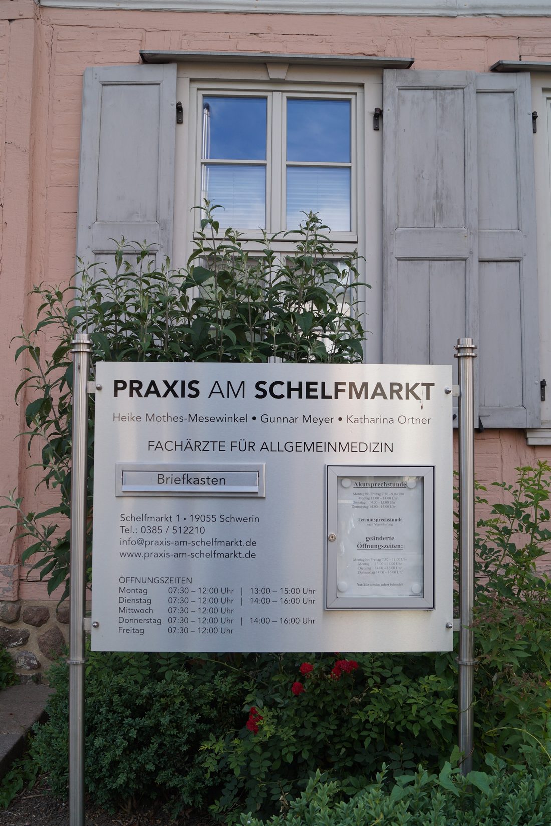 Praxisschild vor dem Eingang mit Fensterläden und Rosen